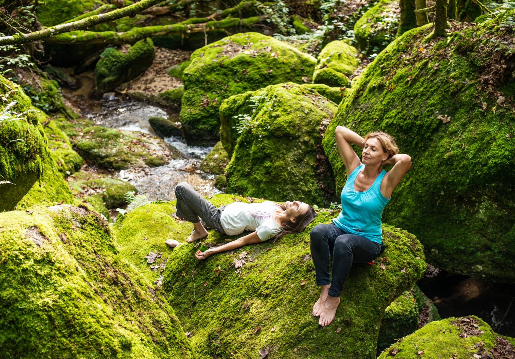 Zwei Frauen sitzen im Wald auf einem Felsen und atmen die Waldluft ein